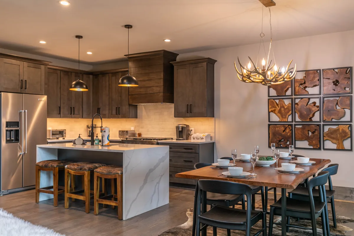 A warm, inviting kitchen and dining area featuring a spacious island, adjacent dining table, decorative wall art, and hanging light fixtures. The scene captures the communal atmosphere ideal for family meals, celebrations, and sharing recipes.