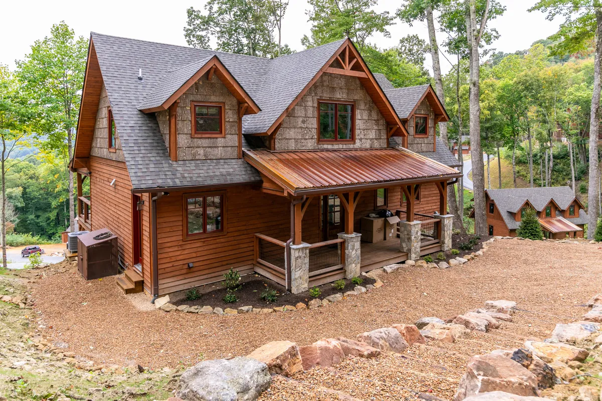 The image displays the front of the wooden cabin, featuring stone pillars, a welcoming porch area, and lush greenery. The porch is set up as an inviting gathering place, perfect for group relaxation, casual meals, or simply enjoying the outdoors. The scene radiates warmth and hospitality, inviting guests to gather and unwind.