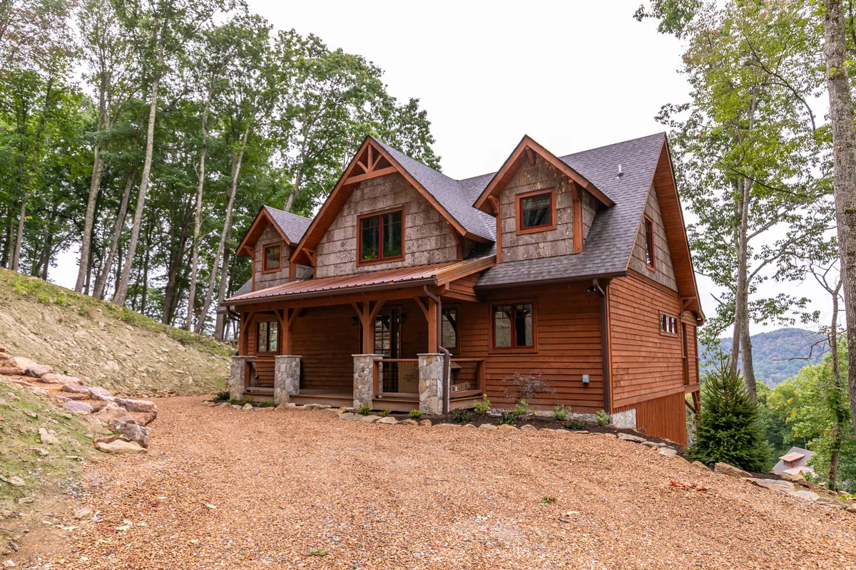 A welcoming exterior view of the wooden cabin, featuring a cozy porch with stone columns and surrounded by lush trees. The setting evokes the tranquility of mountain life and hints at the variety of outdoor experiences available throughout the year.