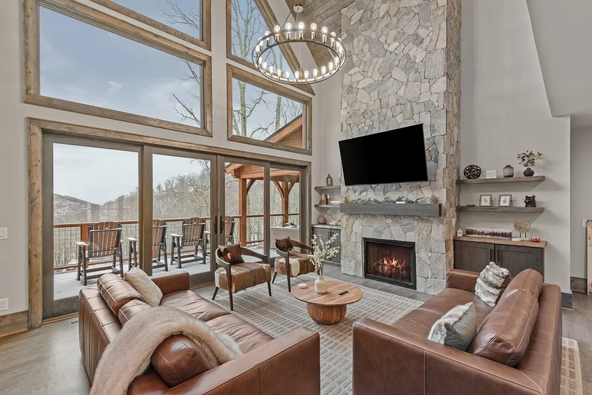 A spacious living area with large windows framing beautiful mountain views. The room features a stone fireplace, chic chandelier, and leather sofas arranged for both group conversations and relaxation. Sunlight pours in, creating a warm and inviting atmosphere ideal for gatherings.