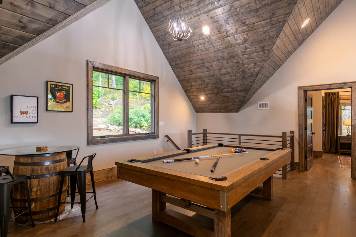An inviting interior game area featuring a pool table and a cozy barrel table set near a large window, allowing natural light to pour in and offering glimpses of the outdoors. The mood is friendly and relaxed, ideal for unwinding after a day outside.