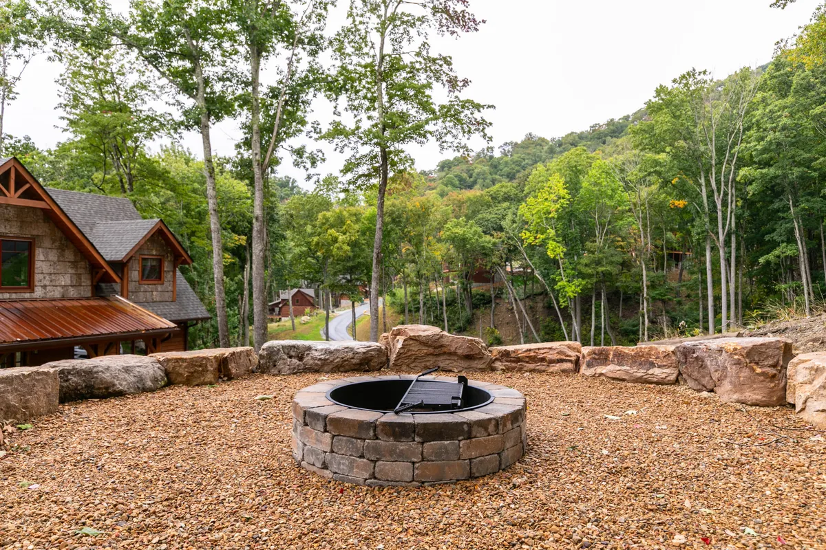 A cozy outdoor fire pit surrounded by natural rocks and tall trees, with a cabin visible in the background. The scene is set for campfire gatherings, storytelling, and stargazing, evoking the communal and magical evenings described in the narrative.