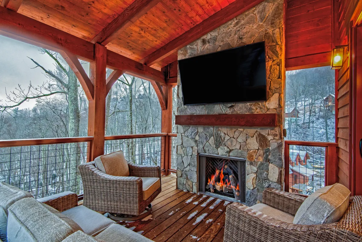This image presents an inviting exterior living space featuring a stone fireplace, comfortable lounge seating, a large TV, and rustic wooden beams. The setting is outdoors, blending natural elements and cozy amenities, ideal for group gatherings and evening entertainment. The warmth of the fire and the view beyond suggest a perfect location for social events.