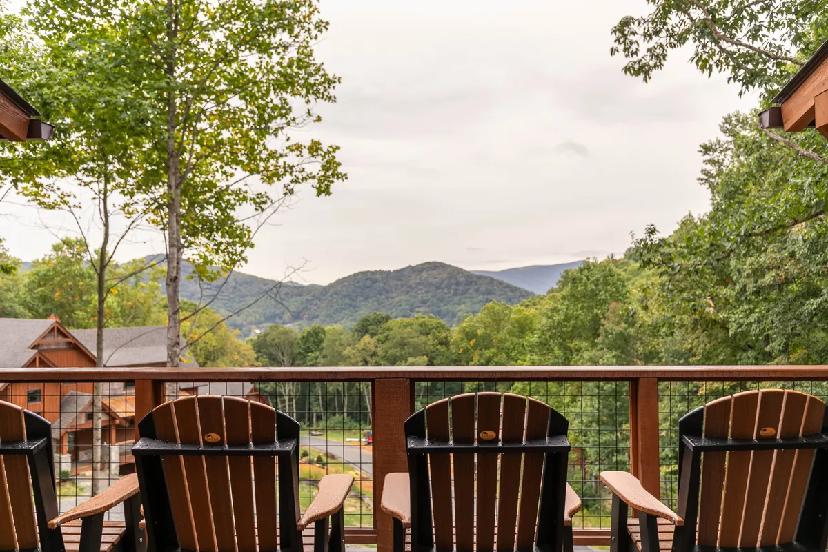 A pair of wooden chairs positioned on a deck facing a stunning mountain view, with lush tree foliage framing the scene. The perspective invites thoughts of quiet reflection, sipping coffee, or enjoying the changing light over the peaks.