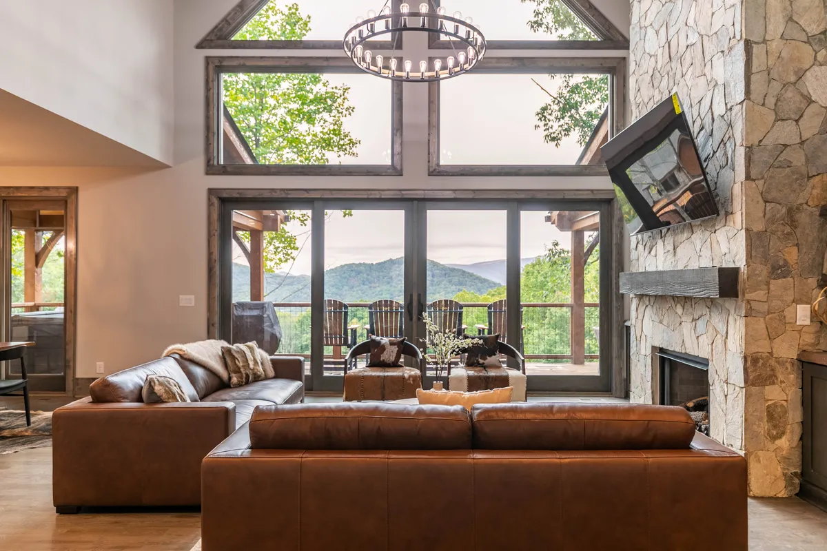 An open-plan living and dining area displaying large windows with mountain views, a stone fireplace, comfortable leather sofas, and a striking chandelier. The arrangement invites both social interaction and quiet relaxation, and the abundance of natural light enhances the cozy atmosphere.