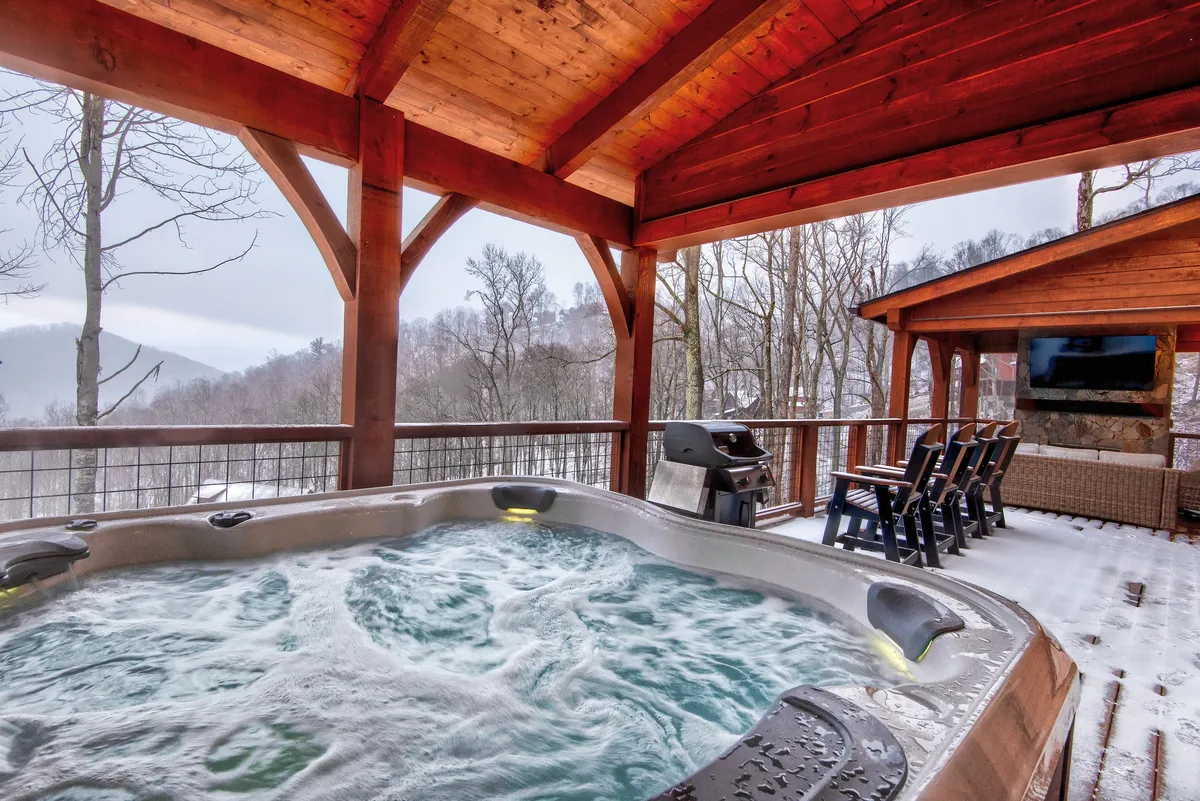 This image features a tranquil wooden deck surrounded by lush trees, with a private hot tub as the central focal point. Comfortable outdoor seating and a grill are also visible, offering an inviting setting for relaxation or social gatherings while enjoying mountain views.
