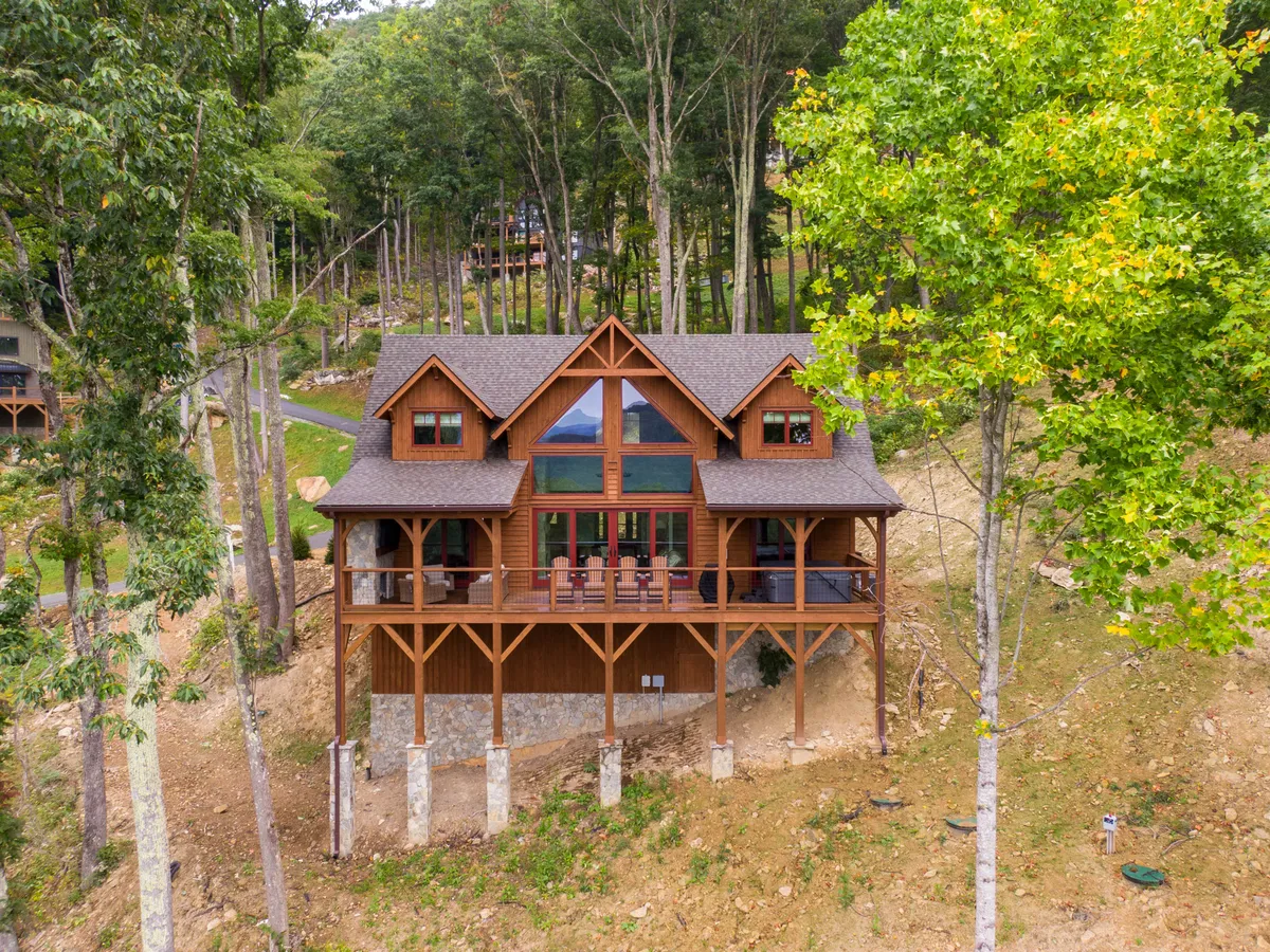 The image showcases the charming wooden exterior of the cabin, with expansive windows inviting natural light and views. The spacious deck, furnished with comfortable chairs, is surrounded by tall trees and opens up to breathtaking sunset vistas. The mood is warm and welcoming, suggesting relaxation and togetherness in a natural mountain setting.