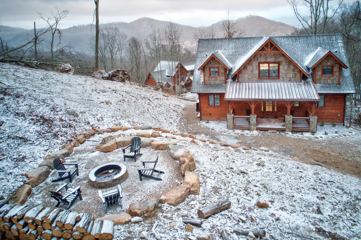 A log cabin house nestled among trees with a prominent fire pit in the foreground, surrounded by comfortable seating. The view opens up to distant mountains, and the setting exudes a cozy, peaceful retreat vibe perfect for outdoor relaxation and stargazing.