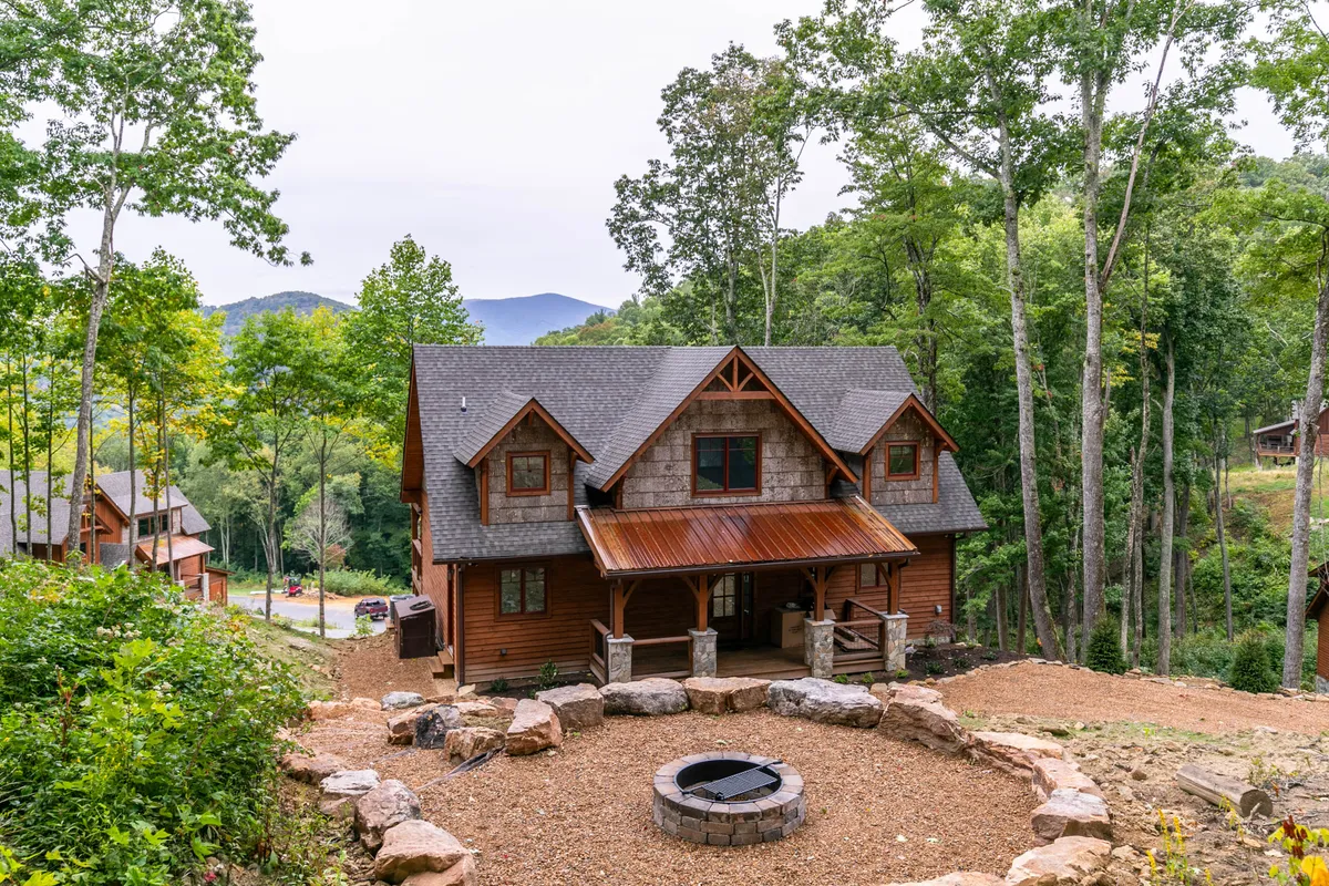 An exterior view highlighting the main entrance, fire pit area, and the house with trees and mountain views in the background. The fire pit is surrounded by seating, creating a cozy gathering space for s’mores and storytelling under the stars.